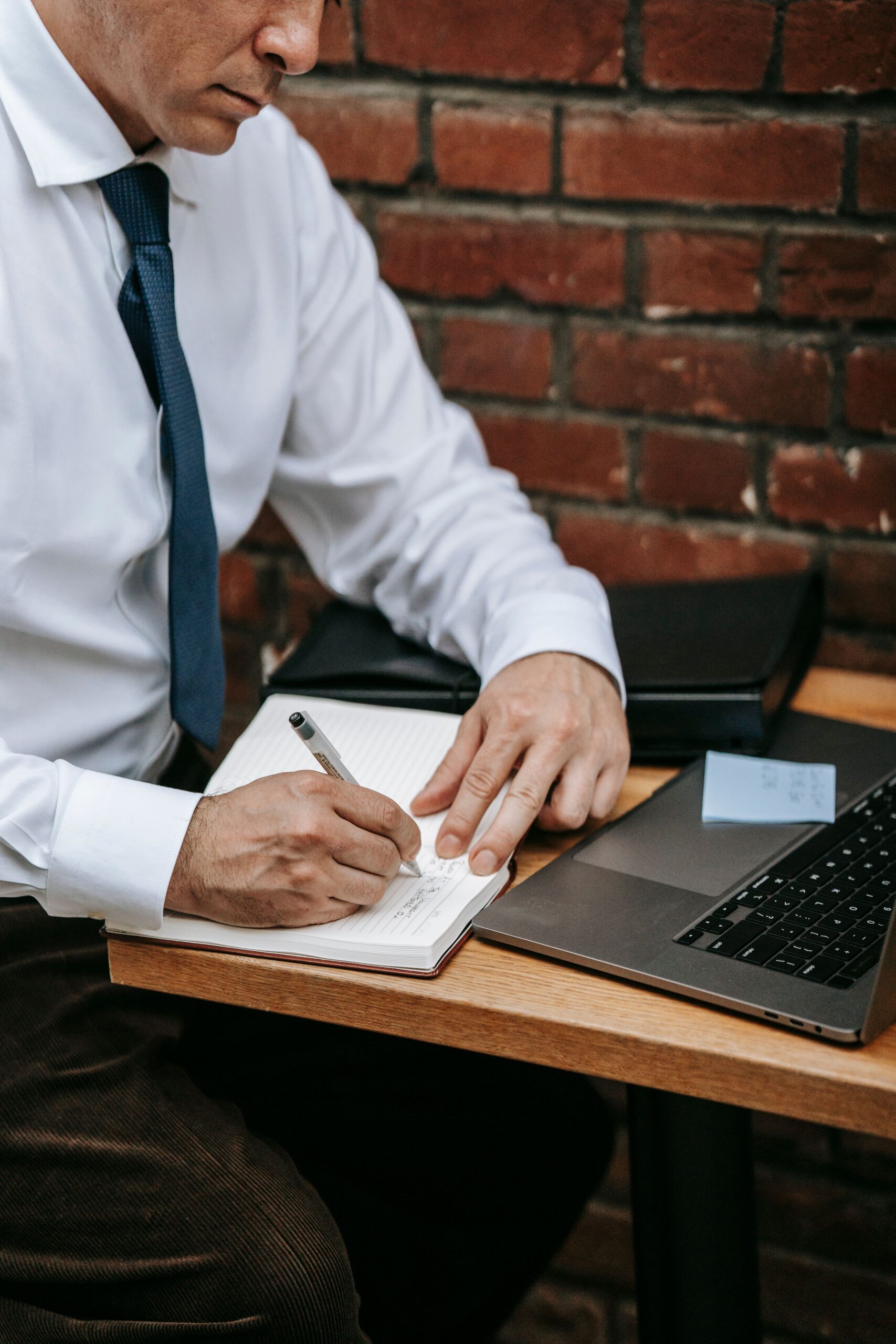 Man in white shirt and tie writing notes at a desk with laptop.